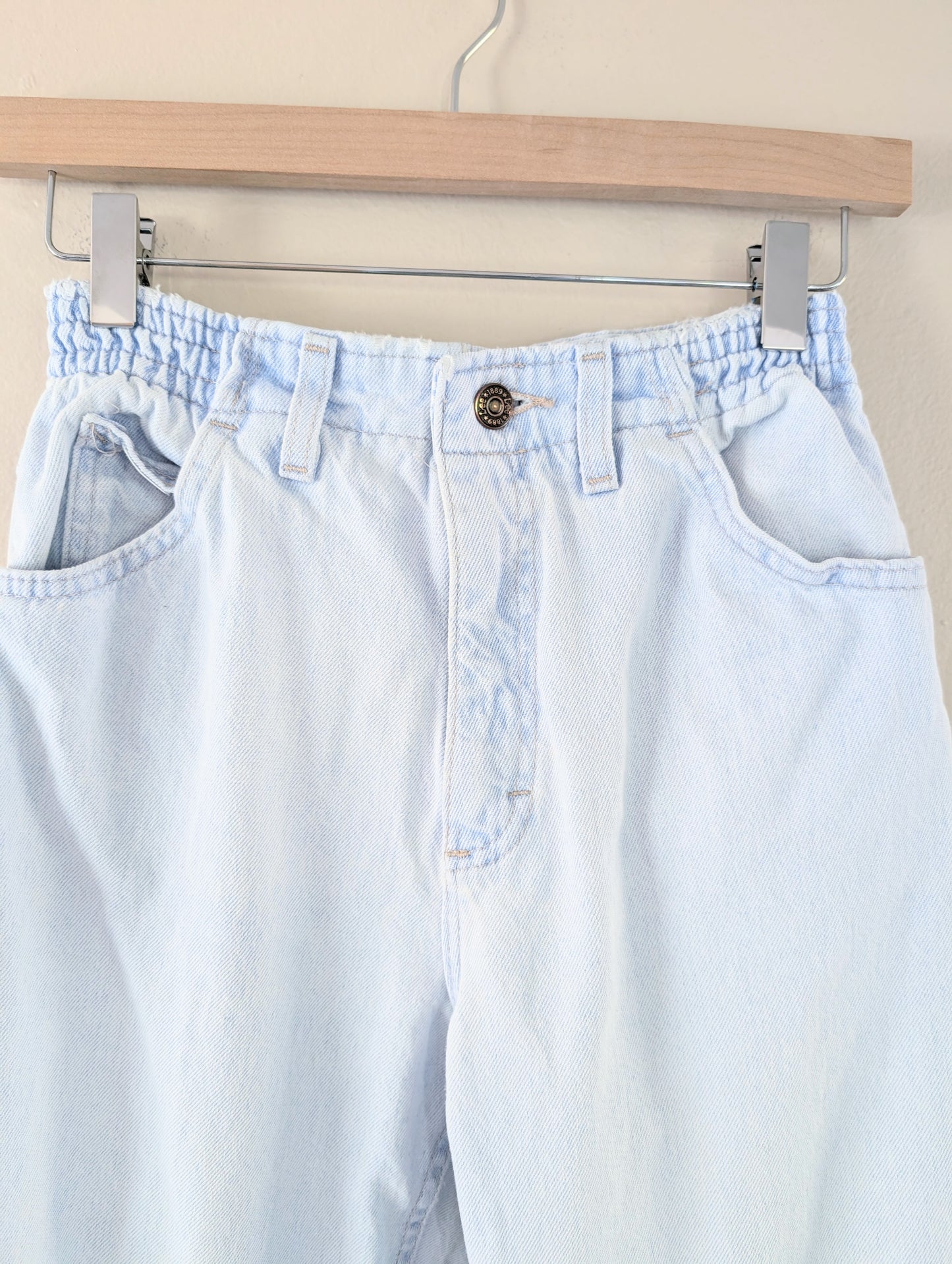 Light blue jeans hanging on a wooden hanger against a beige wall.