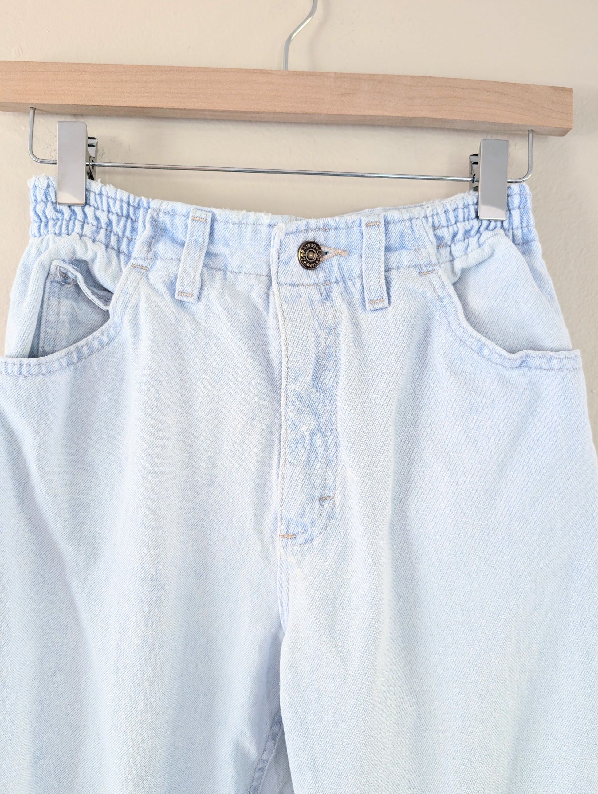 Light blue jeans hanging on a wooden hanger against a beige wall.